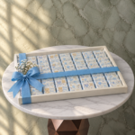 Baby boy chocolate tray amman featuring 100 pieces of blue customized newborn chocolates in a white tray with a blue ribbon and gypsophila flowers.صينية شوكولاتة بيبي بوي في عمان