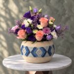 Breeze of elegance amman featuring peach roses, purple lisianthus, and white daisies in a textured ceramic jar.نسمة أناقة