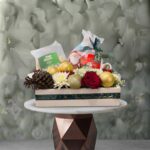 Festive joy christmas hamper amman featuring red roses, white chrysanthemums, pinecones, gold baubles, and holiday gifts in a wooden box.صندوق بهجة العيد