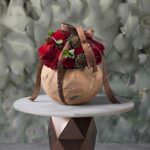Rustic winter charm floral pot amman featuring vibrant red roses, dark chrysanthemums, and pinecones in a round pottery bowl tied with a printed ribbon.