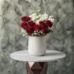 Heartbeat roses amman arrangement featuring vibrant red roses, white stock flowers, and baby's breath in an elegant white ceramic vase.