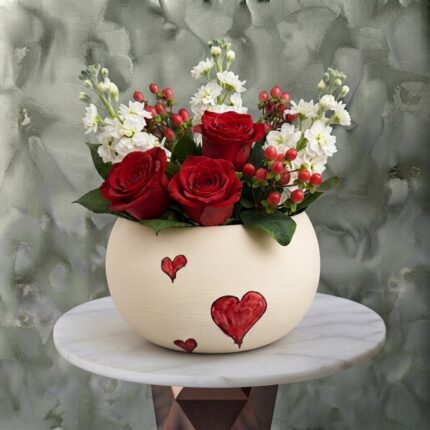 Love whisper arrangement amman featuring fresh red roses, white stock flowers, and red berries in a cream ceramic pot decorated with painted red hearts.