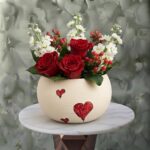 Love whisper arrangement amman featuring fresh red roses, white stock flowers, and red berries in a cream ceramic pot decorated with painted red hearts.