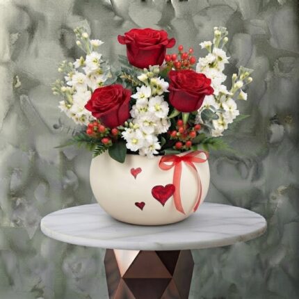 Scarlet valentine amman arrangement featuring vibrant red roses, white stock flowers, and red berries in a cream ceramic pot decorated with red hearts and a ribbon.