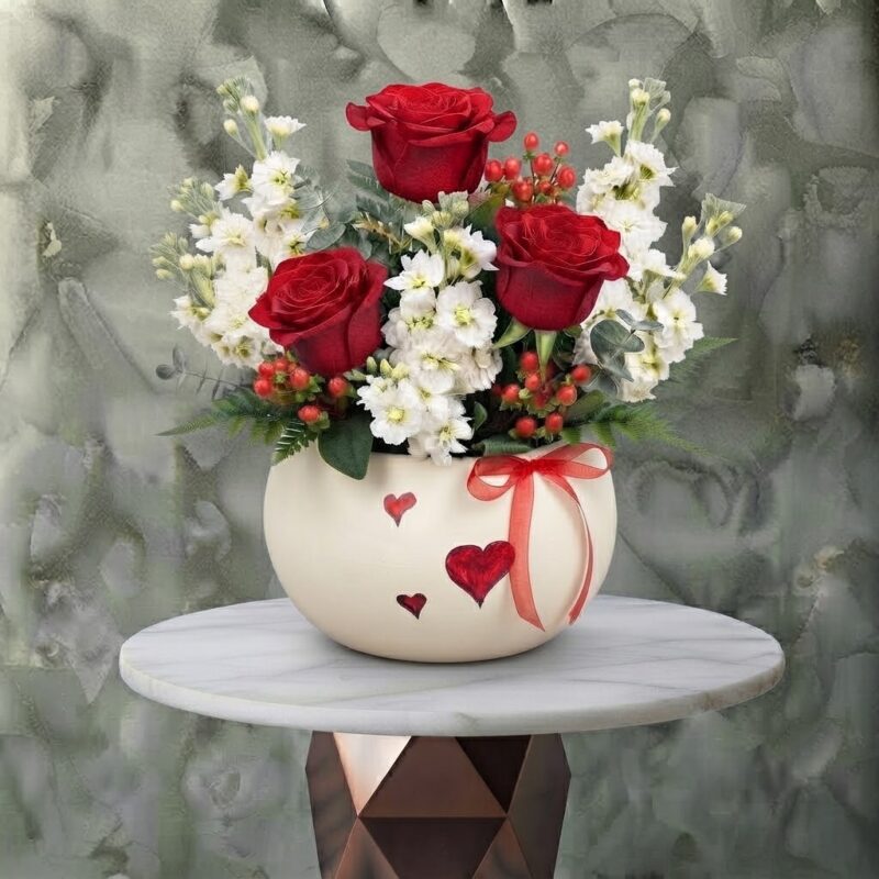 Scarlet valentine amman arrangement featuring vibrant red roses, white stock flowers, and red berries in a cream ceramic pot decorated with red hearts and a ribbon.