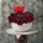 Velvet romance amman arrangement featuring a red teddy bear, rich dark chrysanthemums, roses, and pinecones in a modern white ceramic pot.
