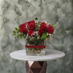 Classic red amman arrangement featuring fresh red roses, white baby's breath, and greenery in a square glass vase tied with a red ribbon.كلاسيك ريد