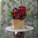 Crimson rustic amman arrangement featuring deep red roses and burgundy blooms with foliage inside a craft paper handle bag.ريف أحمر
