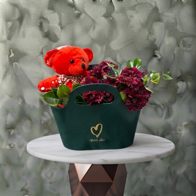 Red emerald amman gift arrangement featuring dark red flowers, eucalyptus, and a plush red teddy bear in a green handle bag.الزمرد الأحمر