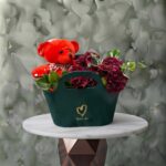 Red emerald amman gift arrangement featuring dark red flowers, eucalyptus, and a plush red teddy bear in a green handle bag.الزمرد الأحمر