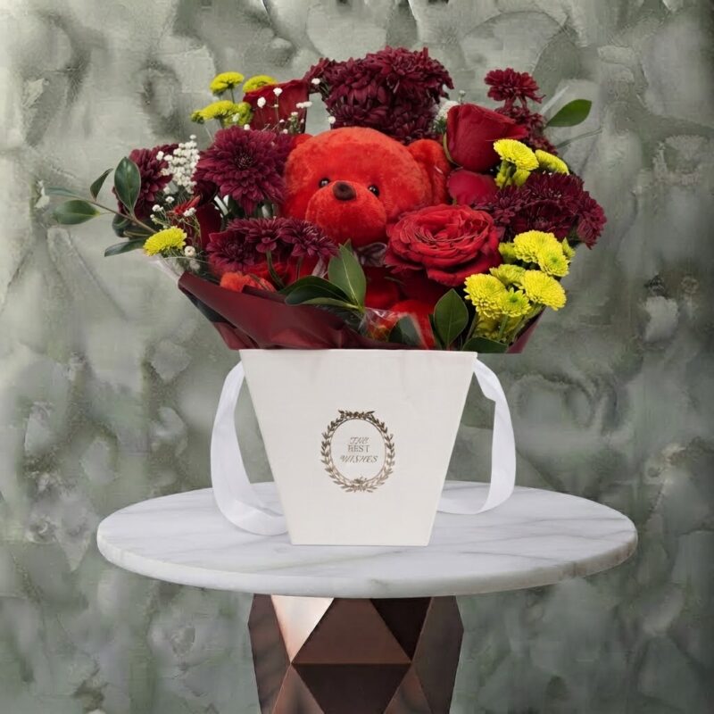 Red trio amman gift arrangement featuring red roses, vibrant seasonal blooms, and a plush teddy bear in a white handle bag.الثلاثية الحمراء