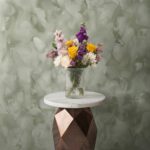 Bright hope vase amman with yellow roses, purple stock flowers, and white chrysanthemums in a clear ribbed glass vase.