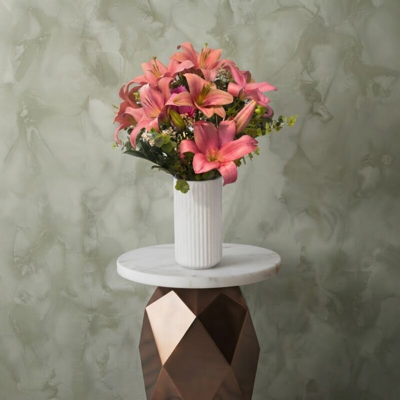 Pink royal lily vase amman with fresh pink lilies and greenery in a white ribbed ceramic vase on a marble stand.