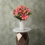 Pink royal lily vase amman with fresh pink lilies and greenery in a white ribbed ceramic vase on a marble stand.