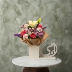 Ummi floral arrangement amman featuring white lily, pink gerbera daisies, and purple statice in a wooden base with Arabic calligraphy escultura.أمي